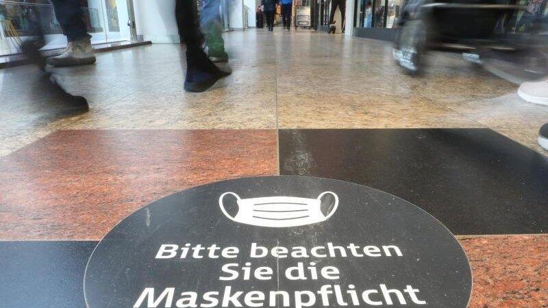 Passanten laufen durch ein Einkaufszentrum auf dessen Fußboden ein Hinweisschild zur Maskenpflicht aufgeklebt ist. Passanten laufen durch ein Einkaufszentrum auf dessen Fußboden ein Hinweisschild zur Maskenpflicht aufgeklebt ist.
